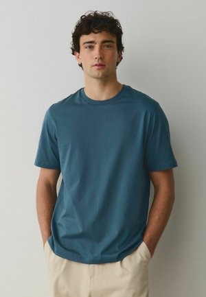 Mladý muž s tmavými kučeravými vlasmi, oblečený v modrej košeli s krátkym rukávom a béžových nohaviciach, stojaci s rukami v kapsách na jednofarebnom pozadí.