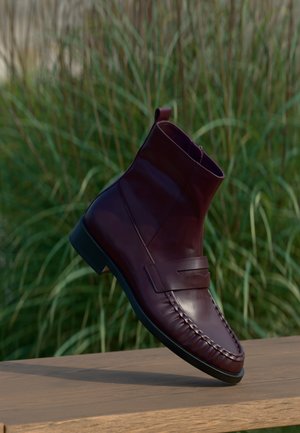 Bottine en cuir marron foncé avec couture moc toe et détail d'une sangle, positionnée sur une surface en bois avec des plantes vertes en arrière-plan.