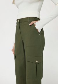 Pantaloni cargo verdi con tasche laterali, frontale piatto e dettagli in metallo argentato. Il tessuto ha una texture liscia e un design strutturato.