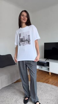 Ženska v široki beli majici s črno-belim potiskom mestnega prizora, sivih hlačah in črnih čevljih stoji v dnevni sobi.