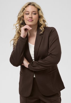Femeie blondă cu păr creț purtând un costum maro și un top alb, sprijinindu-și bărbia în palmă, privind ușor spre o parte pe un fundal simplu.