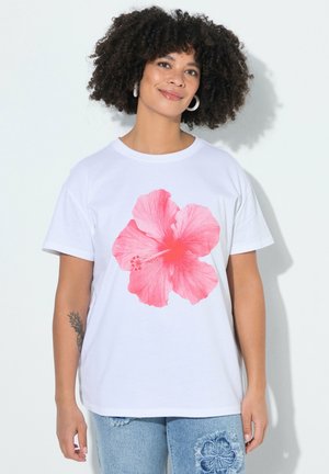 Femme aux cheveux bouclés portant un t-shirt blanc orné d'une grande impression de fleur d'hibiscus rose et un jean bleu avec un motif floral.
