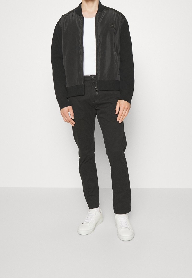 Veste bomber noire avec devant en satin lisse et manches en tricot, associée à un pantalon noir slim et des baskets blanches. T-shirt blanc uni en dessous.