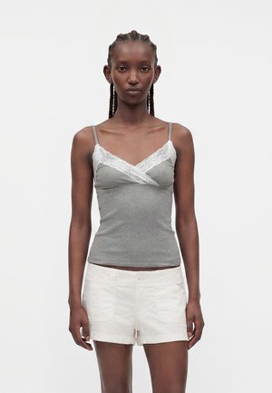 Kvinne med flettet hår har på seg grå camisole med hvit blonderkant og hvite shorts, står foran ensfarget lys bakgrunn.