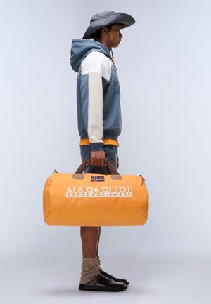 Orange duffle bag with brown leather handles, featuring a white logo, blue and white striped hoodie, gray sun hat, and dark shoes with gray socks.