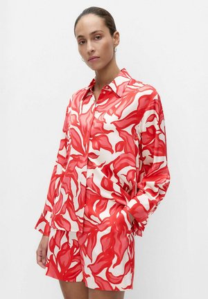Vrouw draagt een rood-wit gemêleerd, gebuttoned langwerpig hemd met bijpassende short, staand met één hand in de zak tegen een witte achtergrond.