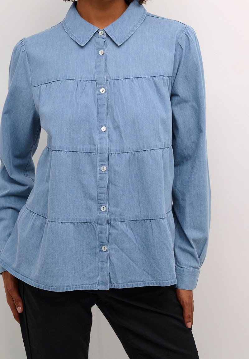 Ljusblå denimskjorta med knappar fram, långa ärmar, krage och horisontella sömdetaljer; har små knappar och en rundad nederkant.