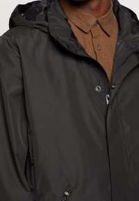 Chaqueta negra ligera con capucha, con cierre de cremallera, botones a presión y bolsillos laterales. Cuello texturizado marrón por debajo.