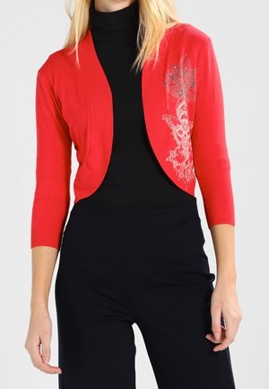 Kobieta ubrana w krótki czerwony sweterek z trójkątnymi rękawami i kwiatowym nadrukiem na czarnym golfie i czarnych spodniach.