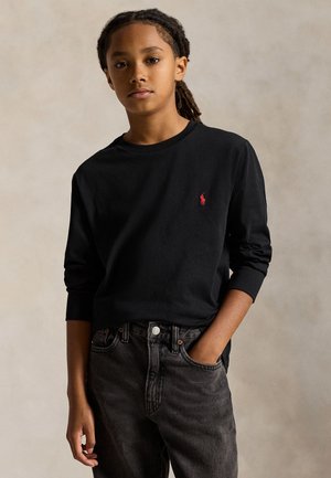 Une jeune personne porte un haut noir à manches longues avec un petit logo rouge sur la poitrine et un jean foncé. Elle se tient devant un fond de studio beige.