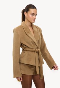 Blazer beige à texture douce, taille ceinturée, col cranté et manches longues, associé à un short assorti. Design minimaliste, sans attaches visibles.