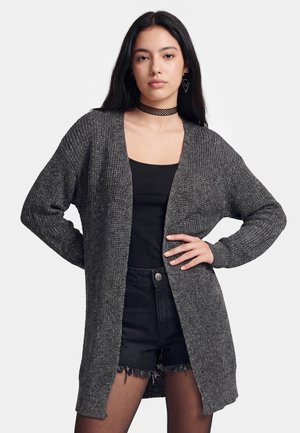 Ung kvinde med langt mørkt hår bærer en mørkegrå strikket cardigan over en sort top og sorte denimshorts med frynset kant.