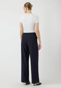 Femme avec les cheveux en chignon portant un t-shirt blanc ajusté, un pantalon noir à taille haute et jambes larges, et des chaussures noires à petits talons, de dos.