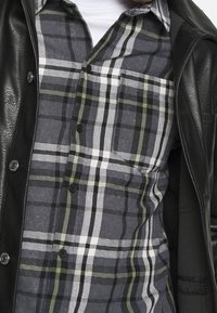 Schwarze Lederjacke über einem grau karierten Hemd. Das Hemd hat grüne, weiße und schwarze Streifen mit einer Knopfverschluss-Design und kurzem Kragen.