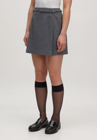 Grijze wollen mini-jurk met een aangesloten taille, riemlussen en plooien. Gecombineerd met zwarte kniekousen en glanzende zwarte loafers.