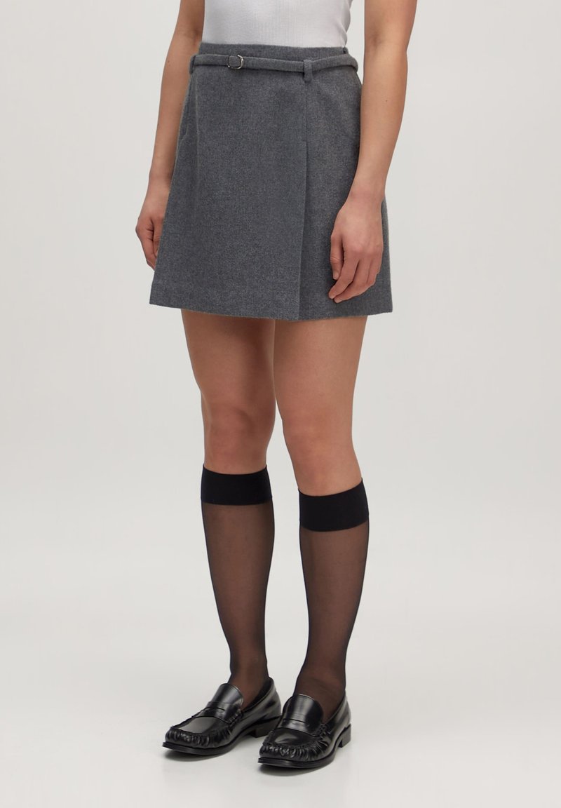 Grijze wollen mini-jurk met een aangesloten taille, riemlussen en plooien. Gecombineerd met zwarte kniekousen en glanzende zwarte loafers.