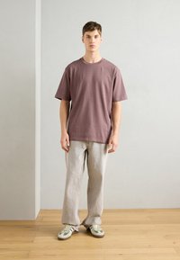 T-shirt de algodão roxa com mangas curtas, combinada com calças cinza claro e ténis com detalhes em verde, contra um fundo neutro.