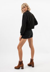 Giacca nera cropped con orlo a coste, abbinata a pantaloni corti neri. Stivaletti in suede marrone con tacco spesso. Modella che volta le spalle, mostrando la vista laterale.