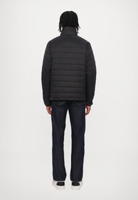 Giubbotto a sacco nero con texture trapuntata e colletto alto, indossato sopra jeans blu scuro e sneakers bianche e nere, visto da dietro.