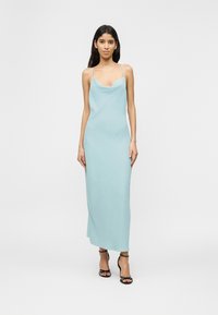 Γυναίκα που στέκεται, φορώντας ένα ανοιχτό μπλε, αμάνικο φόρεμα slip και μαύρα σανδάλια με ψηλό τακούνι και λουράκια, σε λευκό απλό φόντο.