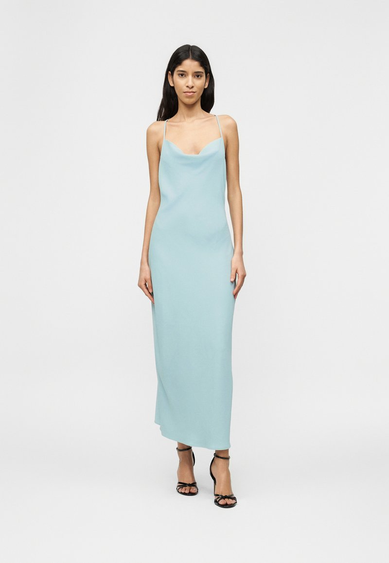 Γυναίκα που στέκεται, φορώντας ένα ανοιχτό μπλε, αμάνικο φόρεμα slip και μαύρα σανδάλια με ψηλό τακούνι και λουράκια, σε λευκό απλό φόντο.