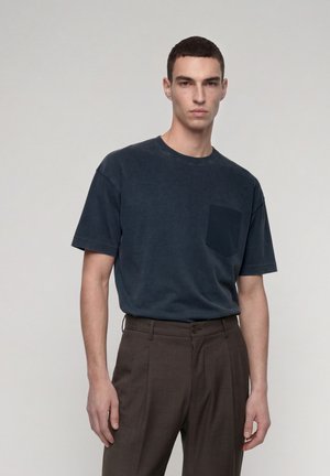 Junger Mann mit kurzem dunklem Haar, der ein dunkelblaues Kurzarmhemd mit Brusttasche und dunkle Hose trägt, steht vor einem einfarbigen Hintergrund.