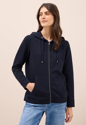 Sweat-shirt zippé marine en tissu doux avec poches avant, capuche à cordon de serrage et poignets côtelés. Associé à un jean bleu clair.