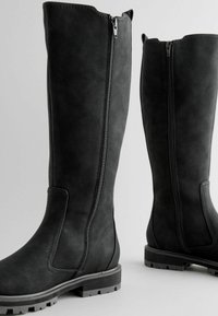 Knee-high black boots made of textured faux leather, featuring a side zipper and pronounced rubber soles with traction detailing.
