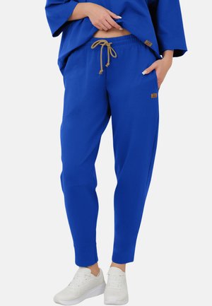 Osoba nosząca królewsko niebieskie spodnie dresowe z beżowym sznurkiem i białe tenisówki, jedną ręką w kieszeni, drugą lekko unoszącą koszulę.