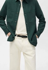 Chemise verte à boutons avec deux poches frontales, portée par-dessus un pull léger côtelé et un pantalon de couleur claire, avec une ceinture noire.