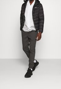 Černá puffer bunda s kapucí, bílá košile s knoflíky a šedými vlnkovými vzory, šedé kalhoty a černé sportovní boty. Jednoduchý, funkční oděv.