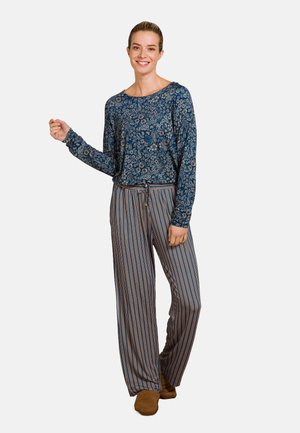 Langlopende blauwe bloemenblouse gecombineerd met gestreepte, brede broek in oranje, blauw en crème, aangevuld met bruine slippers.