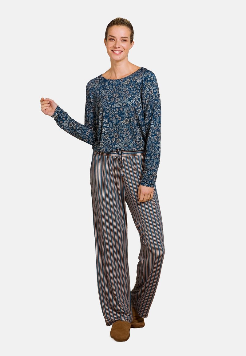 Langlopende blauwe bloemenblouse gecombineerd met gestreepte, brede broek in oranje, blauw en crème, aangevuld met bruine slippers.