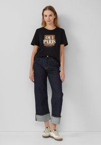T-shirt noir avec le texte "OUI PARIS" sur fond léopard. Associé à un jean tapered en denim noir et des baskets beiges.