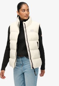 Gilet imbottito crema con collo alto, chiusura con zip, tasche frontali e dettaglio logo sobrio, indossato sopra un maglione nero lavorato a maglia e jeans blu.
