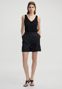 Zwarte V-hals tanktop gecombineerd met hoge zwarte linnen shorts met zijzakken en een opgerolde zoom, gezet tegen een neutrale achtergrond.