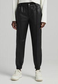 Mustat tekonahkaiset joggers-housut, joissa on sileä pinta, joustava vyötärönauha, resorit ja napitus edessä. Yhdistetty valkoisiin tennareihin.