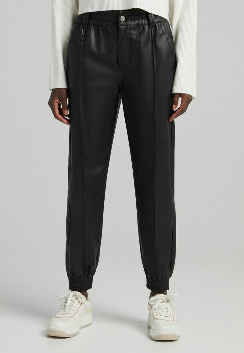 Mustat tekonahkaiset joggers-housut, joissa on sileä pinta, joustava vyötärönauha, resorit ja napitus edessä. Yhdistetty valkoisiin tennareihin.