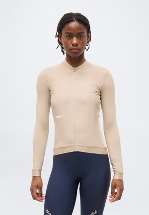 Γυναίκα που φοράει μπεζ αθλητικό τοπ με φερμουάρ και μακριά μανίκια και μπλε σκούρο κολάν με υφή, στέκεται μπροστά σε απλό φωτεινό φόντο.