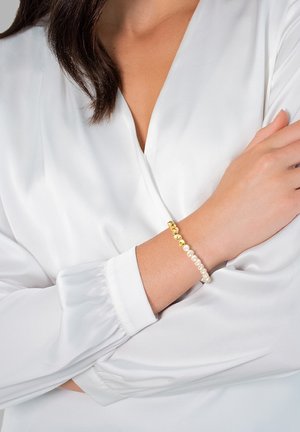 Bracciale in oro e perle caratterizzato da un mix di perle dorate lucide e perle con texture. Indossato su un polso, accanto a una blusa bianca con maniche arricciate.