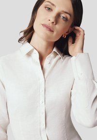 Biała lniana bluzka z guzikowym kołnierzykiem, z długimi rękawami i dopasowanym krojem. Teksturowana tkanina i subtelny połysk.