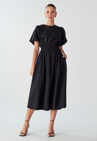 Črna midi obleka s kratkimi napihnjenimi rokavi in elastičnim pasom. Gladek material z stranskimi žepe in zračna A-linija silhueta.