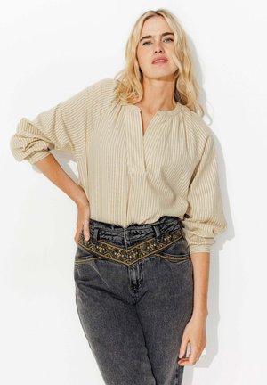 Femme blonde portant un chemisier beige à rayures et un jean noir taille haute avec broderies florales, debout avec une main sur la hanche.