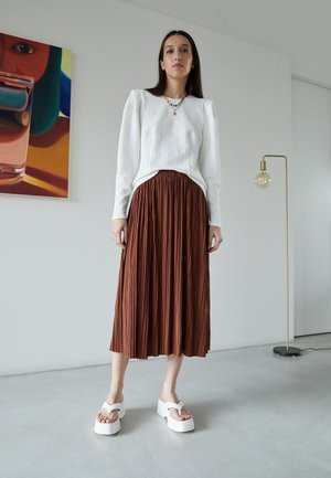 Haut à manches longues texturé blanc, jupe maxi plissée marron et sandales compensées blanches. Design minimaliste avec une silhouette épurée et des détails subtils.