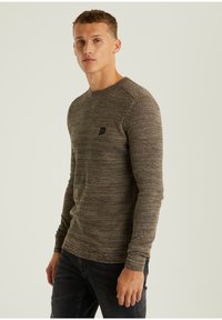 Langärmliger Pullover in einem strukturierten, braunen Strickmuster mit einer figurbetonten Silhouette und einem kleinen schwarzen Logo auf der Brust.