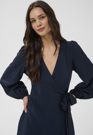 Hosszú sötét hajú nő, mély V-nyakú, tengerészkék átlapolt ruhában, melynek puffos hosszú ujjai vannak, kezét csípőjén tartva áll.