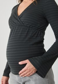 Crna majica dugih rukava s V-izrezom i flared rukavima; ima suptilnu rebrastu teksturu i nabranu tkaninu ispod grudnog koša. Pouch omogućava prilagodbu za trudnički trbuh.