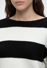 Cou et bas du visage d’une femme portant un pull en maille rayé noir et blanc avec un col rond et des cheveux foncés détachés.