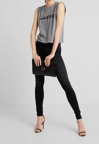 Top sin mangas gris acortado con el logo "GUESS", combinado con jeans ajustados negros. Los accesorios incluyen un bolso negro texturizado con un detalle de anillo dorado.