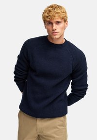 Marineblauer, gerippter Strickpullover mit Rundhalsausschnitt und langen Ärmeln, versehen mit strukturiertem Stoff und taillierten Bündchen, kombiniert mit hellen Khakihosen.